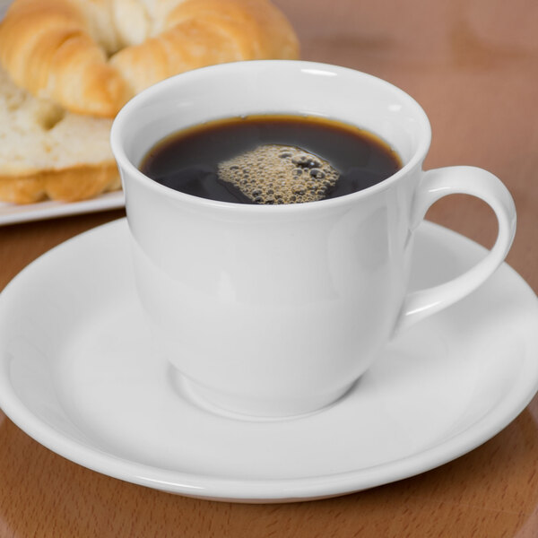 A white porcelain cup filled with black coffee sits on a matching saucer, with a croissant in the background.