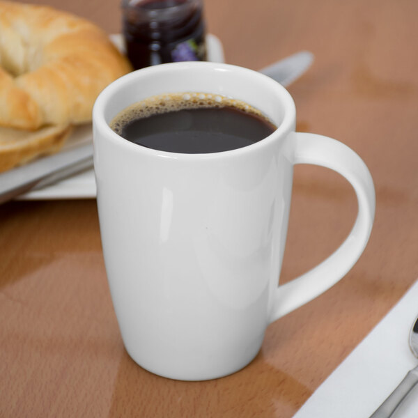 A Libbey white porcelain mug filled with coffee sitting on a table with a croissant.