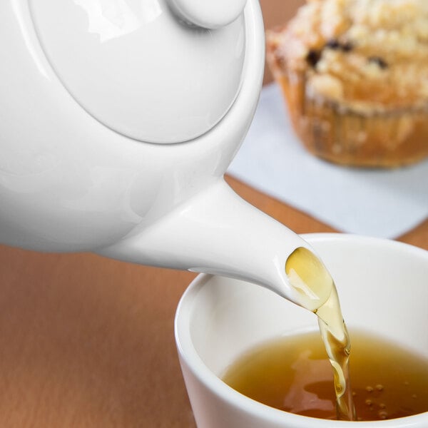A white teapot is pouring tea into a white cup, with a muffin in the background.
