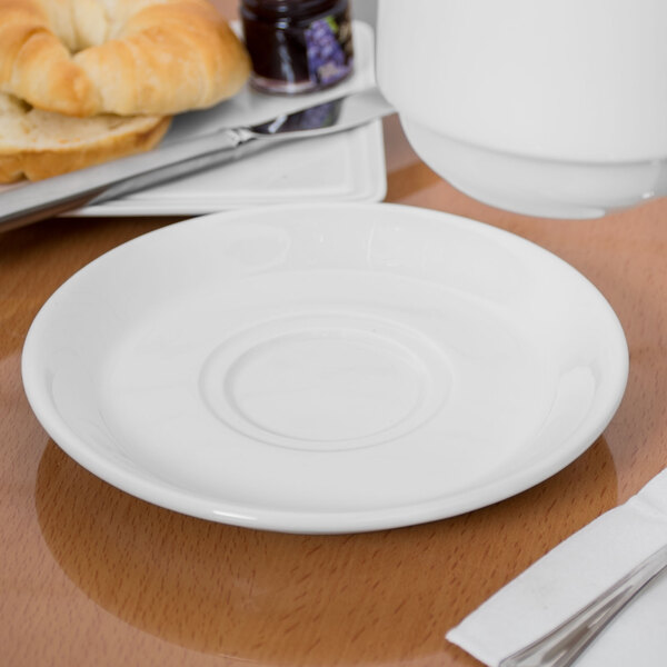 A Libbey white porcelain tea saucer on a table with a cup of coffee and a croissant.