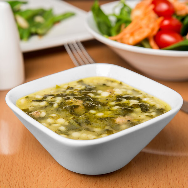 A Libbey Slenda white square porcelain bowl filled with soup and a bowl of salad on a table.