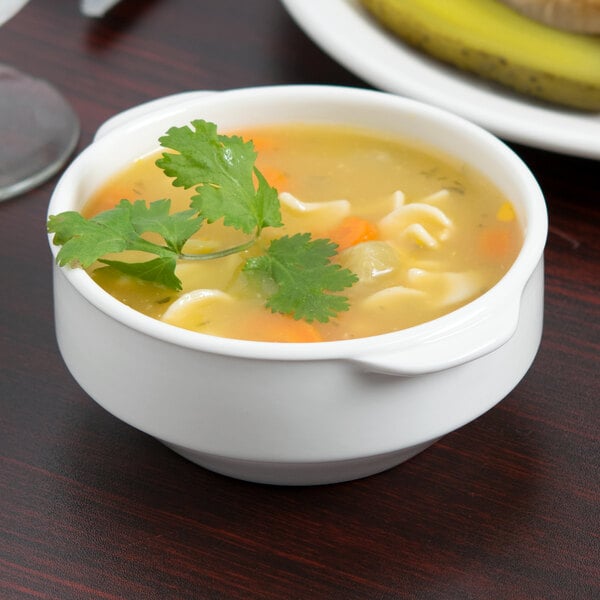 A Libbey white porcelain soup cup with two handles filled with soup and a sandwich on a table.