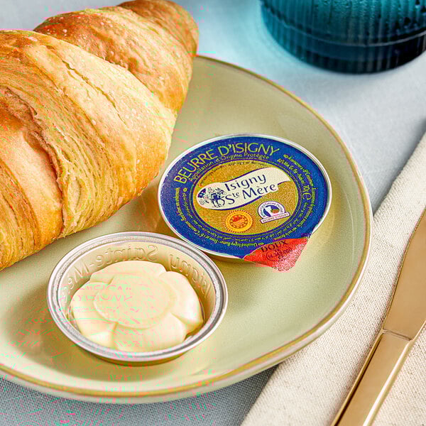 A plate with a croissant and Isigny Sainte-Mere unsalted butter cup on it.