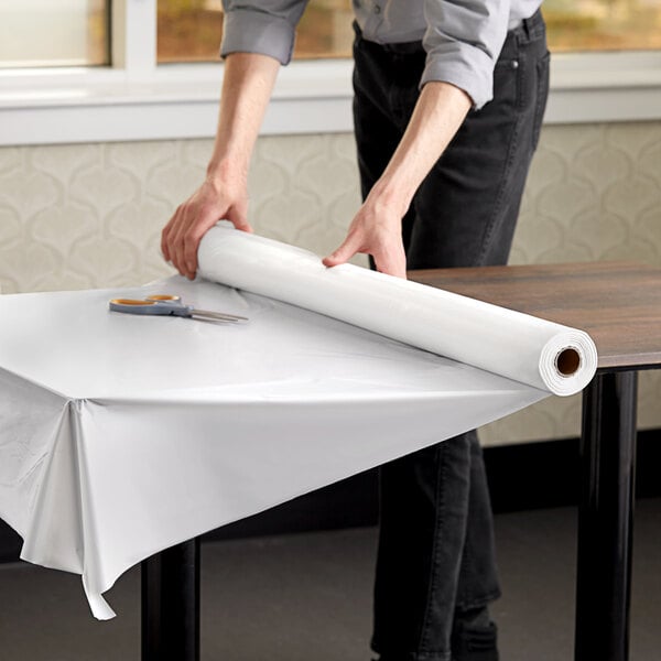 A person rolling a Choice white plastic table cover roll onto a table.