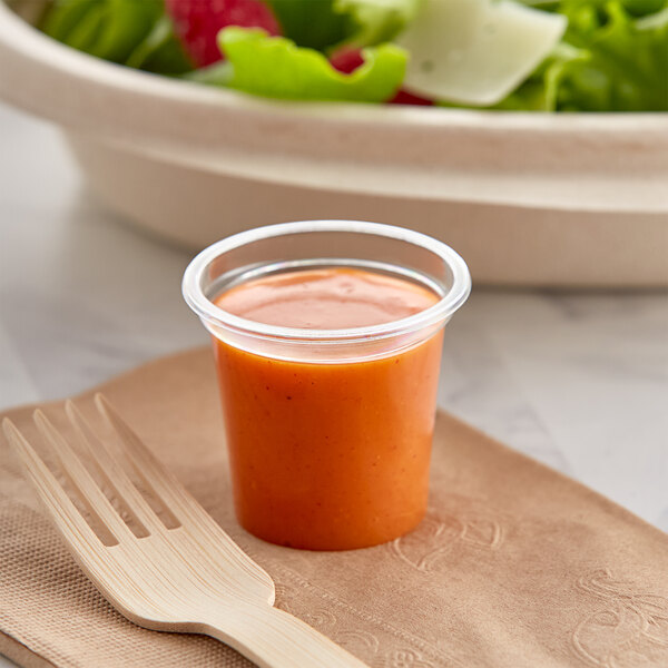 A World Centric compostable plastic cup of orange sauce next to a wooden fork.