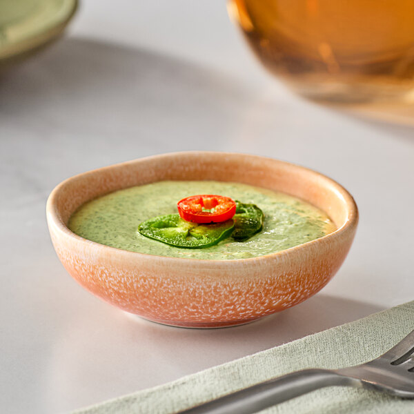 A bowl of green sauce with a red pepper on top in an Acopa Primrose Peach sauce cup.