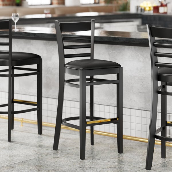 Three Lancaster Table & Seating black wood ladder back bar stools with black vinyl cushions sit in front of a bar.