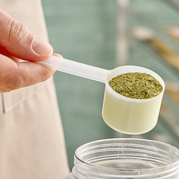 A person holding a 53 cc polypropylene scoop full of green powder over a plastic container.