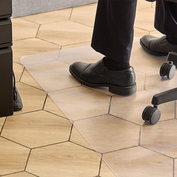 A person wearing black shoes standing on a 360 Office Furniture rectangular clear chair mat on a wooden floor.