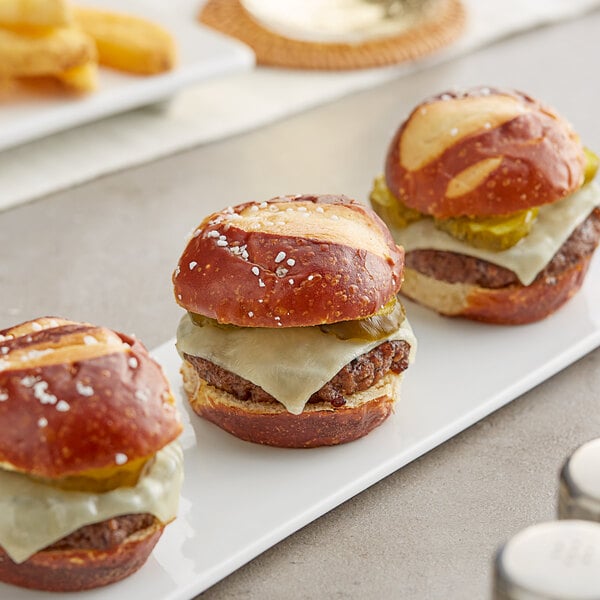 A row of King's Hawaiian Frozen Pretzel Slider Rolls on a white plate with hamburgers.