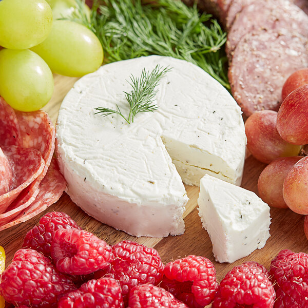 A piece of Cypress Grove PsycheDillic goat cheese on a wooden surface with a slice cut out, grapes, and other fruit.