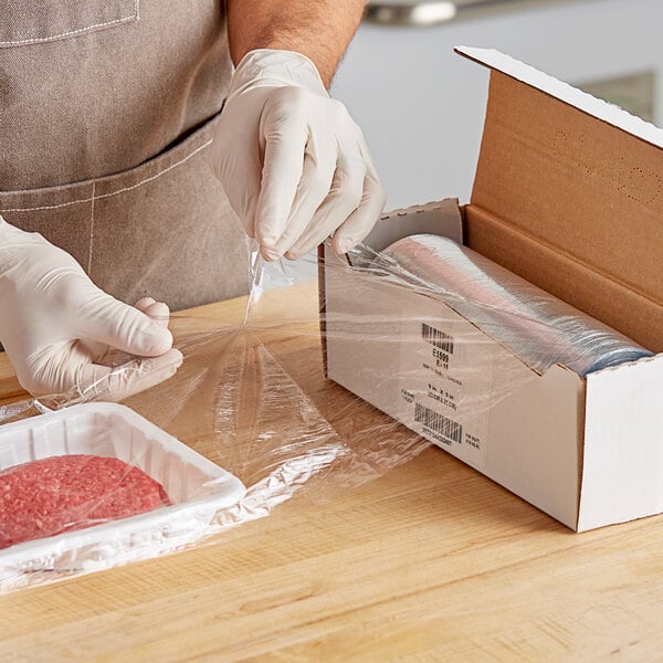 A person wearing gloves and holding Anchor Packaging 9" x 9" perforated plastic wrap over a pile of ground meat on a counter.