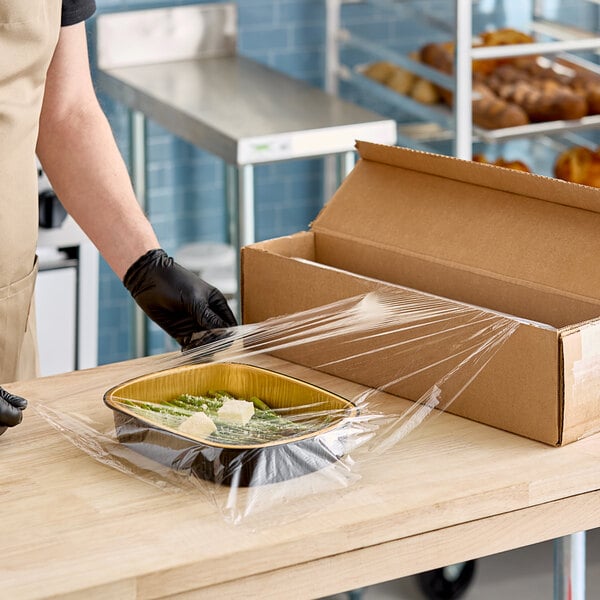 A person wearing black gloves using Anchor Packaging perforated plastic wrap to cover a dish of food.