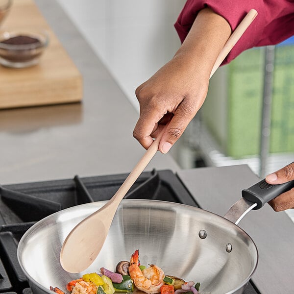 A person cooking food in a pan on a stove with a Choice 18" wooden spoon.