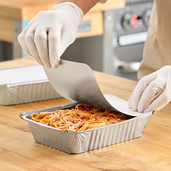 A person wearing gloves using a spatula to put food in a Western Plastics foil container.