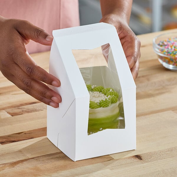 A person holding a Southern Champion white bakery box with a green cupcake inside.