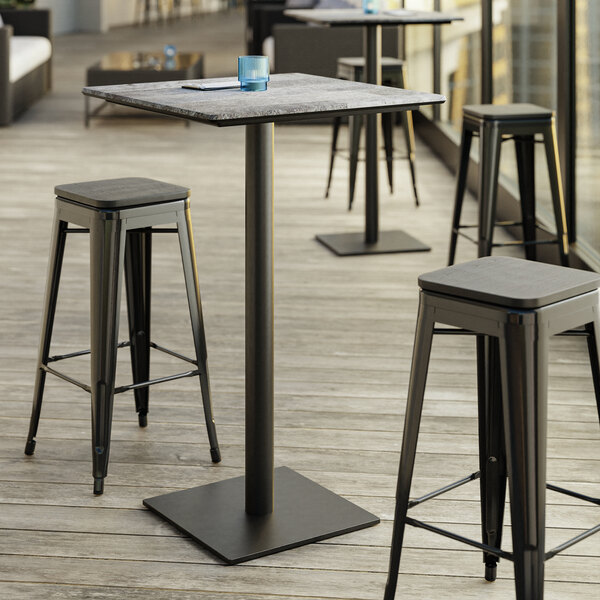 A Lancaster Table & Seating Excalibur bar height table with a textured Toscano finish and square base plate with stools on a patio.