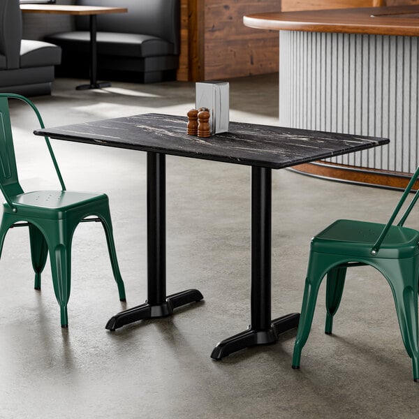 A rectangular table with a dark, marble-like top and black double pedestal base, accompanied by green metal chairs in a modern indoor setting.