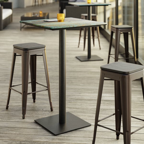 A Lancaster Table & Seating bar height table with a textured metal finish and square base plate with stools on a patio.