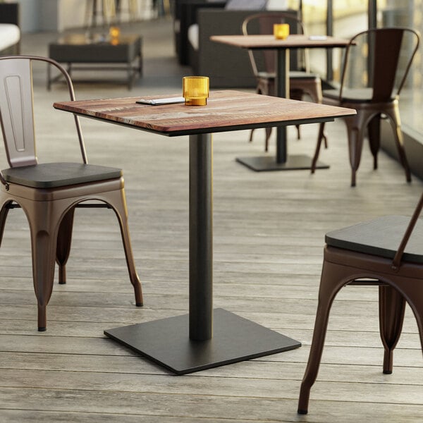 A square wooden table with a black metal pedestal base, surrounded by metal chairs on a wooden deck, with a small yellow candle holder on top.