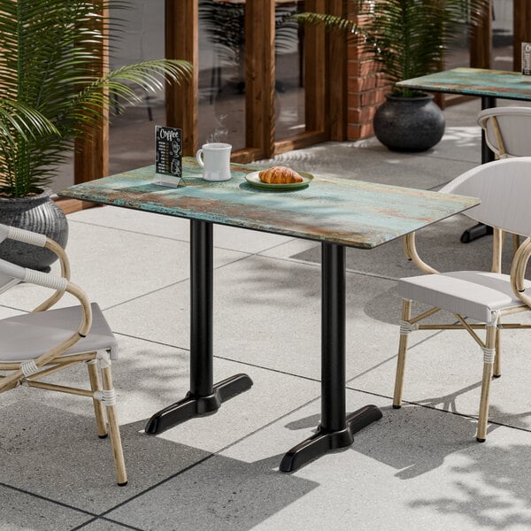 A rectangular outdoor table with a weathered, textured surface and black metal legs, set with a cup, plate, and menu card, surrounded by chairs on a patio.