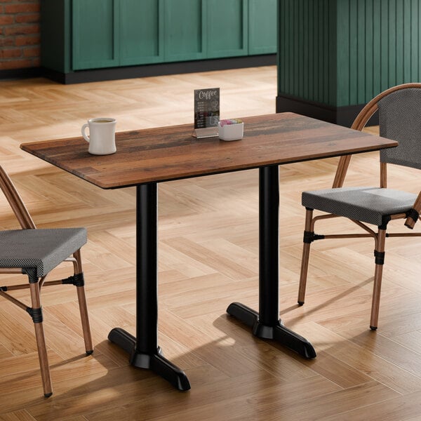A rectangular table with a wood-like textured top and two black metal pedestal legs, accompanied by two chairs in a cafe setting.