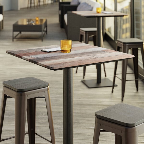A Lancaster Table & Seating square table top with a textured wooden finish on a table with stools and a glass on it.