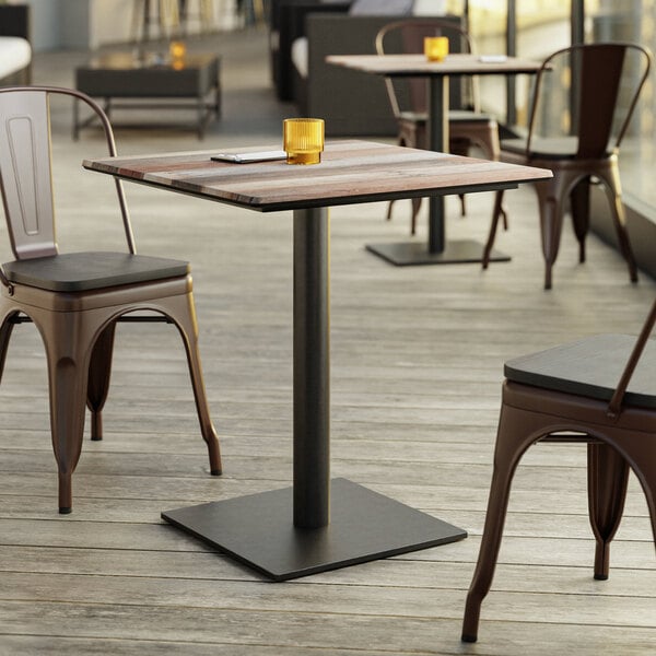 A square table with a mixed wood plank top and a black metal pedestal base, surrounded by metal chairs on a wooden deck.