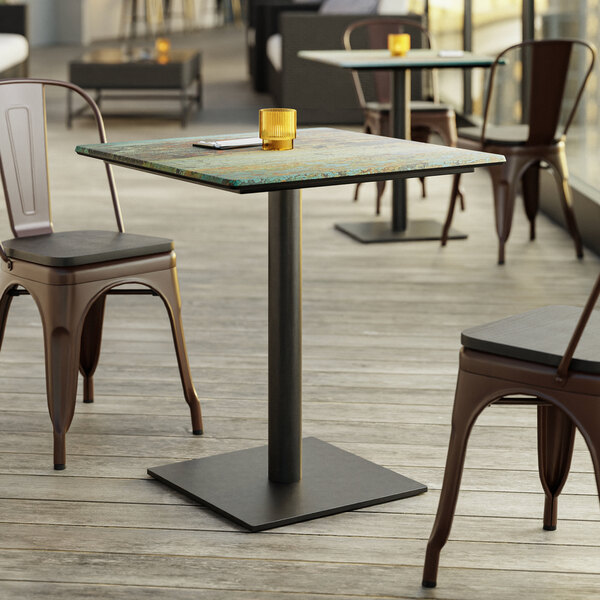 A square table with a textured, weathered-looking top and a black metal pedestal base, surrounded by metal chairs on a wooden deck.
