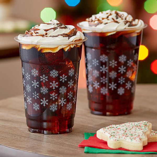 Two Choice Snowflake PET plastic cold cups filled with brown liquid with whipped cream and ice on a table with cookies.