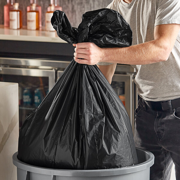 A man putting a Trinity Plastics black trash bag in a trash can.