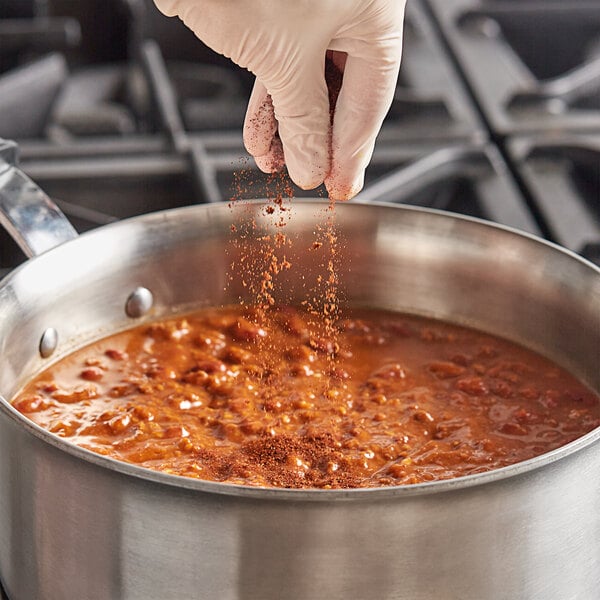 A gloved hand pouring Badia Chili Powder into a pot of food.