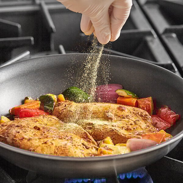 A gloved hand sprinkling Badia Adobo Seasoning on chicken and vegetables in a pan.