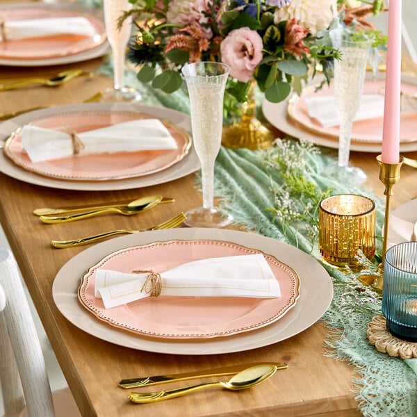 A beautifully set dining table featuring blush pink plates with gold rims, gold cutlery, white napkins tied with twine, glassware, candles, and a floral centerpiece.