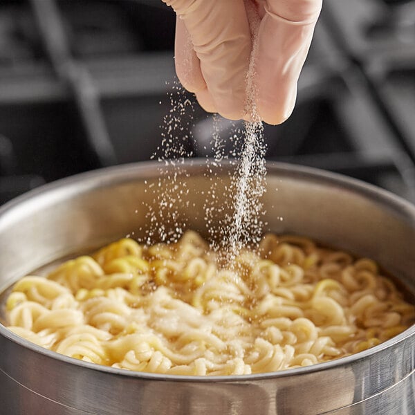 A gloved hand pouring Badia MSG powder into a bowl of noodles.