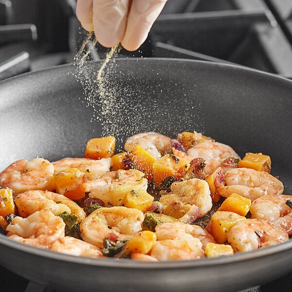 A hand sprinkling Badia Adobo Seasoning on shrimp in a skillet.