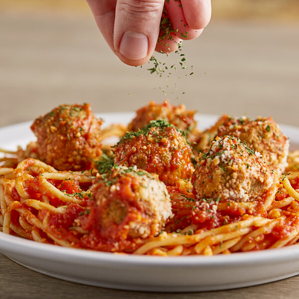 A hand sprinkling Badia Parsley Flakes over a plate of spaghetti and meatballs.