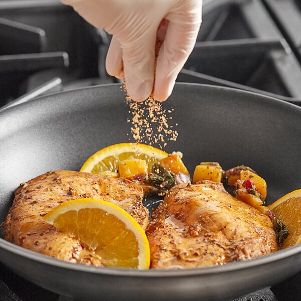 A person's hand sprinkling Badia Orange Pepper on chicken and oranges in a pan.