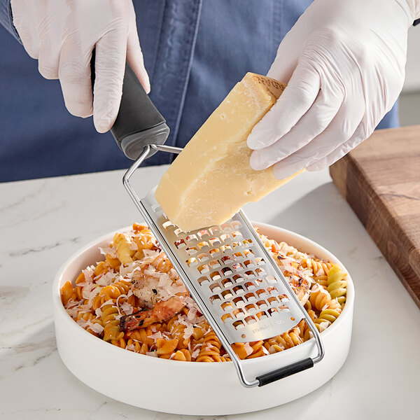 A handheld extra coarse paddle grater being used to grate cheese over a bowl of pasta.