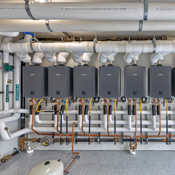 A row of Rinnai SENSEI CXP160iN wall-mounted tankless water heaters connected to various pipes and valves in a utility room.
