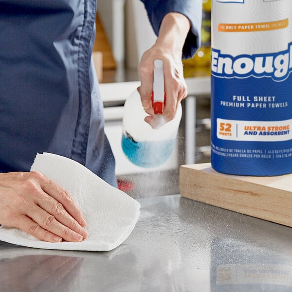 A person cleaning a table in a professional kitchen with Lavex paper towels sprayed with a bottle.