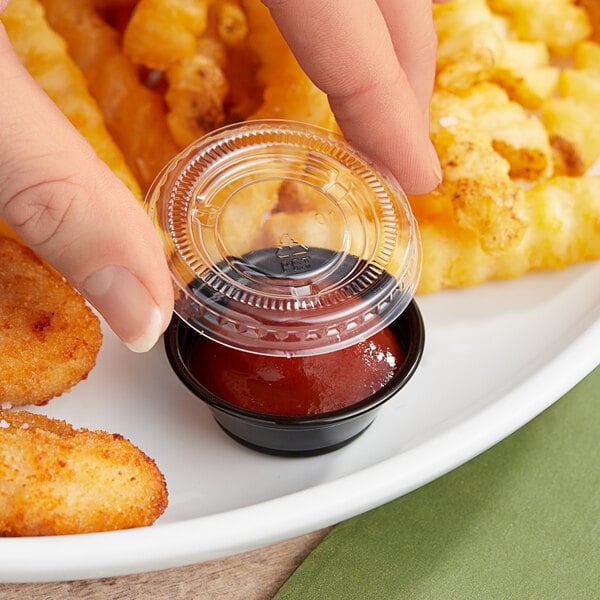 A hand holding a Choice black plastic souffle cup of red liquid over a plate of fried chicken.
