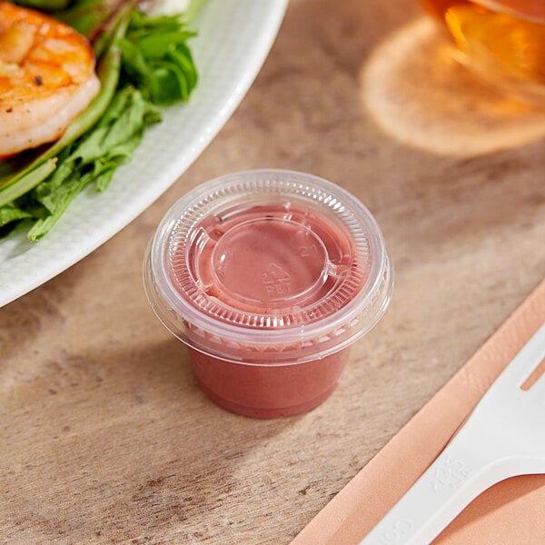 A plastic container of food with a clear plastic lid next to a plate of food with a white spoon and a cup of pink sauce.