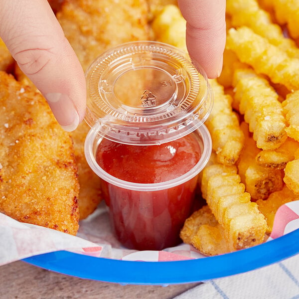 A hand holding a Choice clear plastic souffle cup with red sauce on a plate of chicken nuggets.