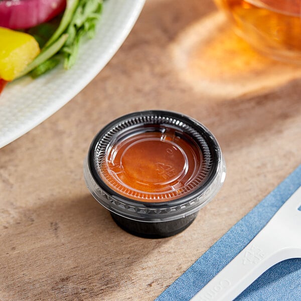 A black plastic souffle cup with brown liquid inside next to a plate of salad.