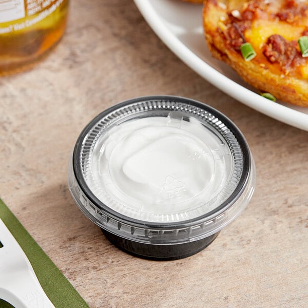 A black Choice plastic souffle cup with white lid filled with white cream next to a plate of food.