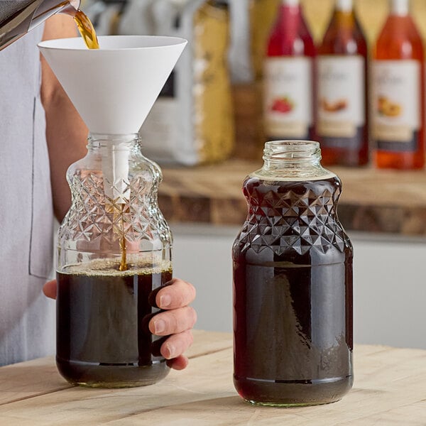 A person pouring dark liquid into a clear glass bottle with a white cap.