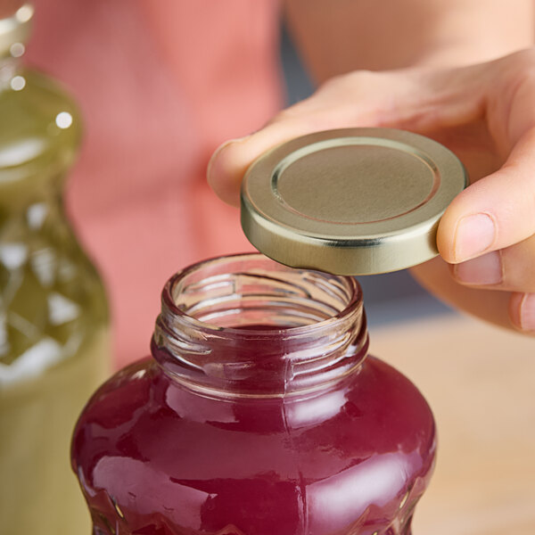 A person holding a 48 Lug gold metal lid over a jar of red liquid.
