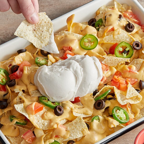 A person holding a tortilla chip dipped in WayFare vegan sour cream on a bowl of nachos.