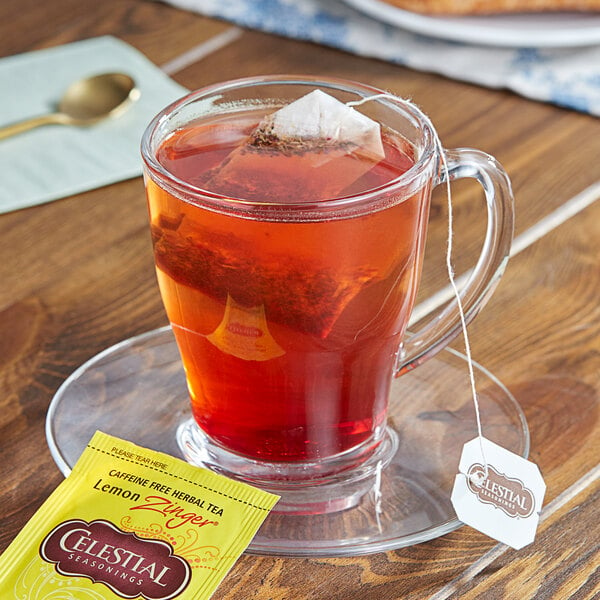 A glass cup of Celestial Seasonings Lemon Zinger tea with a tea bag on a saucer.